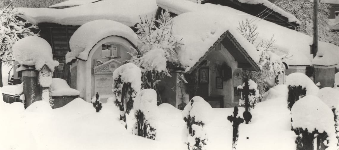 Schwarzweißfoto: Alter Friedhof in Mayrhofen im Winter – Grabkreuze ragen aus hohem Schnee; dahinter kleine Kapelle mit Rundbogen, Häuser und steile Berghänge.