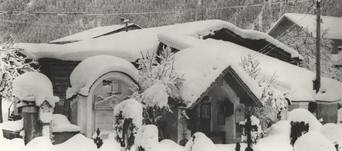 Black-and-white photograph: Old cemetery in Mayrhofen in winter – grave crosses protrude from deep snow; behind them, a small chapel with a round arch, houses and steep mountain slopes.