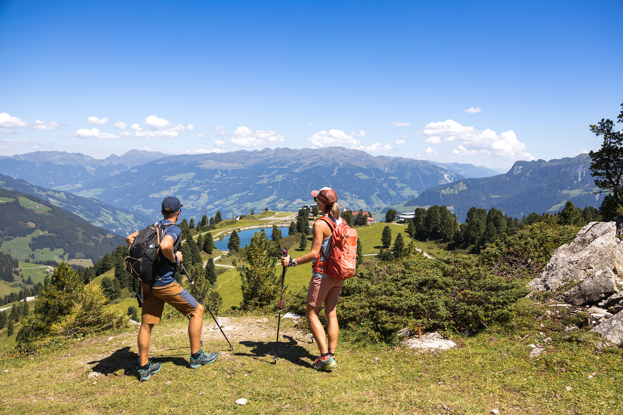 Two hikers with backpacks and hiking poles stand on a grassy hilltop on the Penken in the Zillertal valley. Before them stretches a panorama of mountains, forests, and blue sky.
