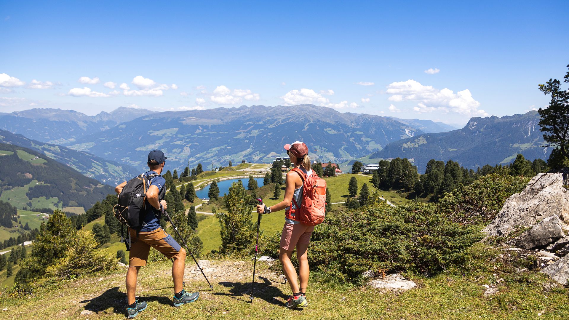 Zwei Wandernde mit Rucksäcken und Wanderstöcken stehen auf einer grasbewachsenen Anhöhe am Penken im Zillertal. Vor ihnen erstreckt sich ein Panorama aus Bergen, Wäldern und blauem Himmel.