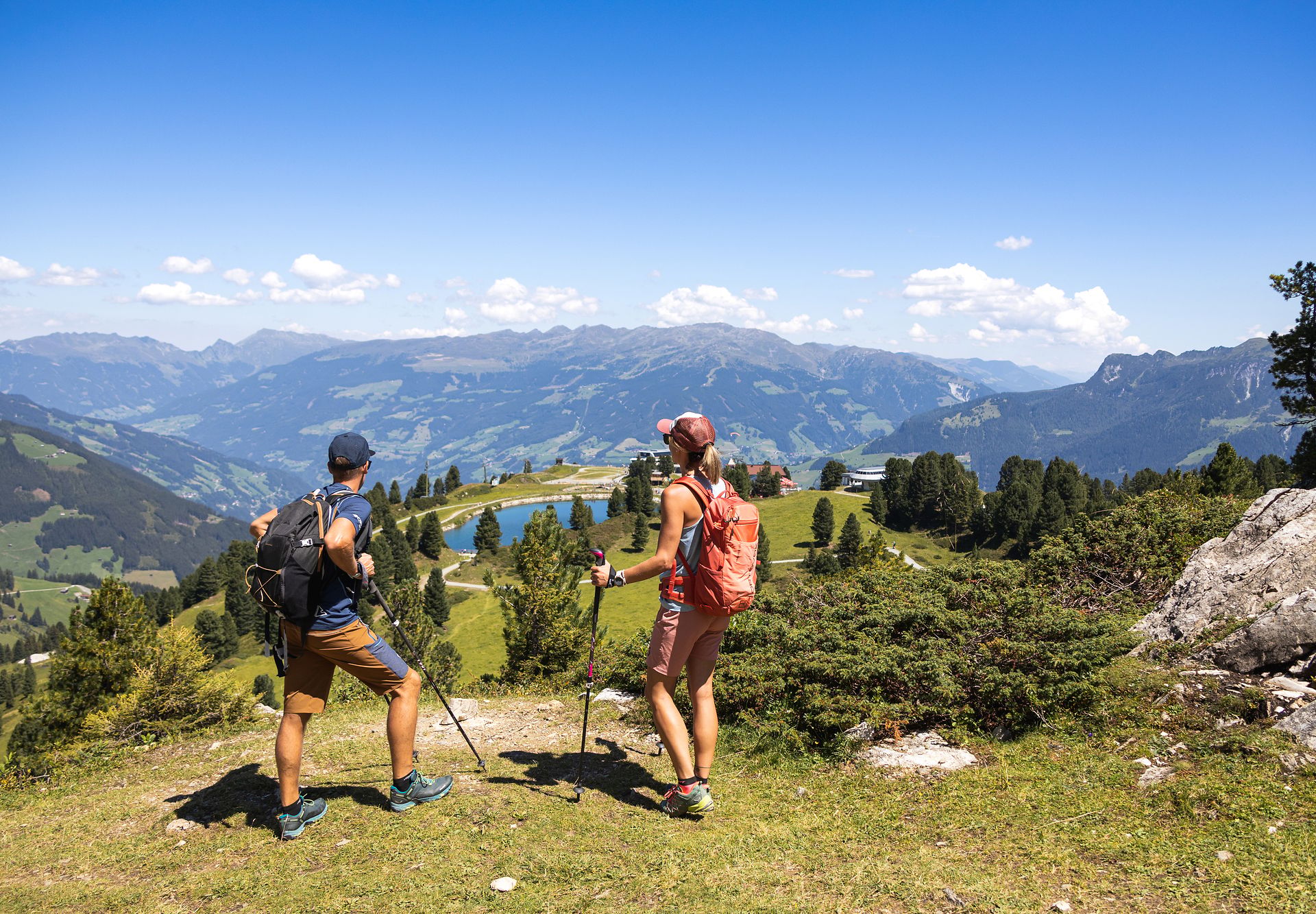 Zwei Wandernde mit Rucksäcken und Wanderstöcken stehen auf einer grasbewachsenen Anhöhe am Penken im Zillertal. Vor ihnen erstreckt sich ein Panorama aus Bergen, Wäldern und blauem Himmel.