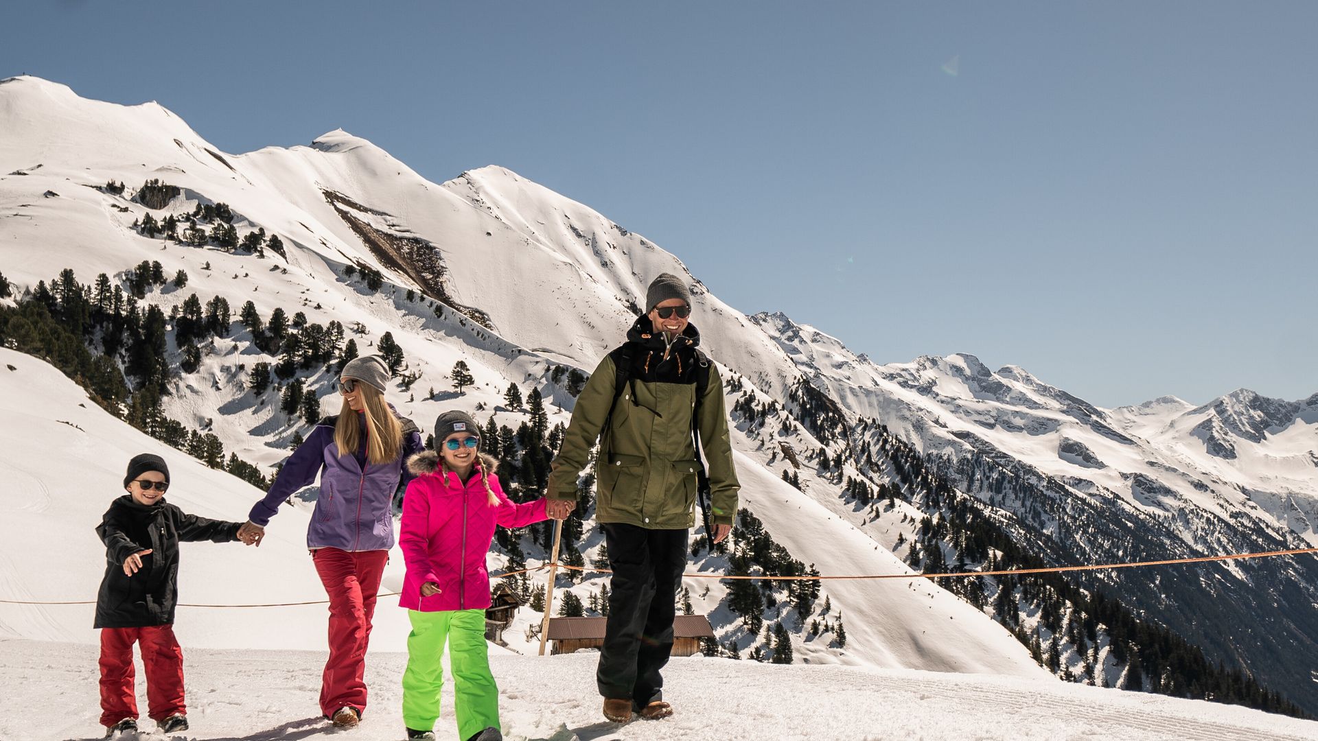 A family enjoys winter hiking on Mount Ahorn in Mayrhofen, walking along a sunny groomed trail with views of the Zillertal Alps in the background.