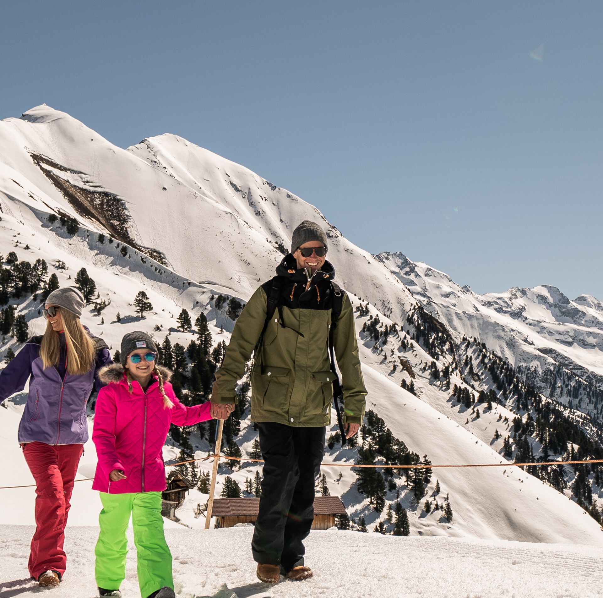 A family enjoys winter hiking on Mount Ahorn in Mayrhofen, walking along a sunny groomed trail with views of the Zillertal Alps in the background.