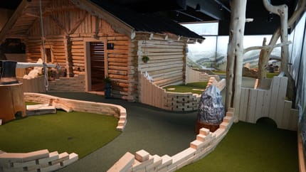 Das Bild zeigt eine Indoor-Adventure-Golf-Anlage mit alpinem Thema. Im Vordergrund sind Bahnen mit grünem Kunstrasen und Holzbegrenzungen zu sehen. Zentral steht eine große Holzhütte im Blockhausstil, daneben ein künstlicher Felsen und Baumstämme.
