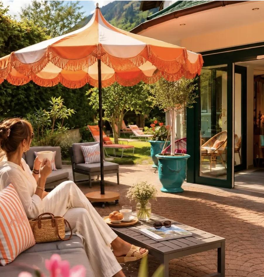 Woman relaxing outdoors under an orange and white striped umbrella, drinking coffee, with garden furniture and plants nearby.