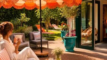 Woman relaxing outdoors under an orange and white striped umbrella, drinking coffee, with garden furniture and plants nearby.