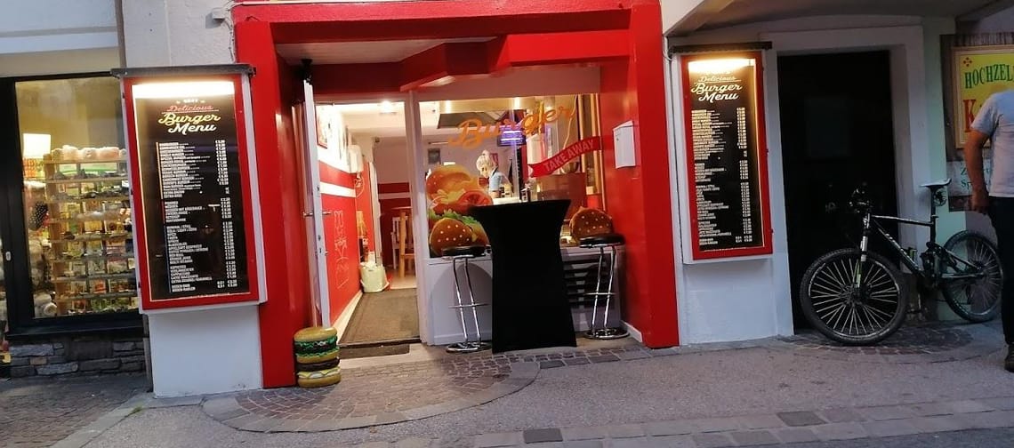Das Bild zeigt die Burger Station von außen. Der Eingangsbereich ist in Rot gehalten und wirkt klein und kompakt. Die Szene vermittelt den Eindruck eines gemütlichen, schnellen Imbisslokals.