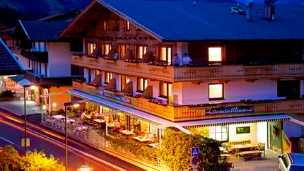 The picture shows Gasthof Zillertaler Weinstadl at night. The wooden building is atmospherically lit, standing out against the dark sky. A car is parked in the lot, while the silhouettes of the mountains in the background are faintly visible.