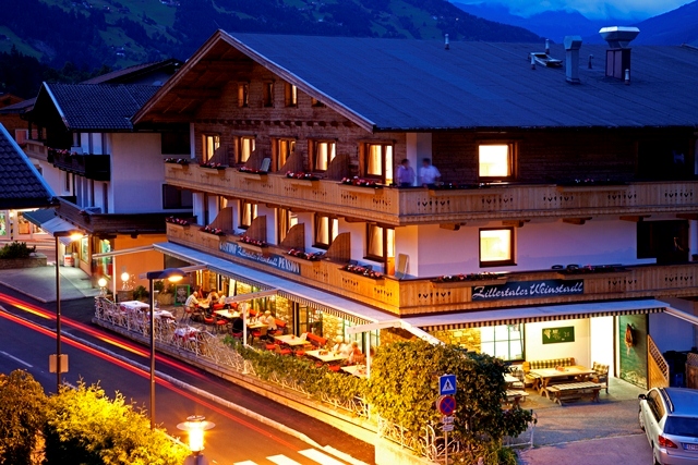 The picture shows Gasthof Zillertaler Weinstadl at night. The wooden building is atmospherically lit, standing out against the dark sky. A car is parked in the lot, while the silhouettes of the mountains in the background are faintly visible.