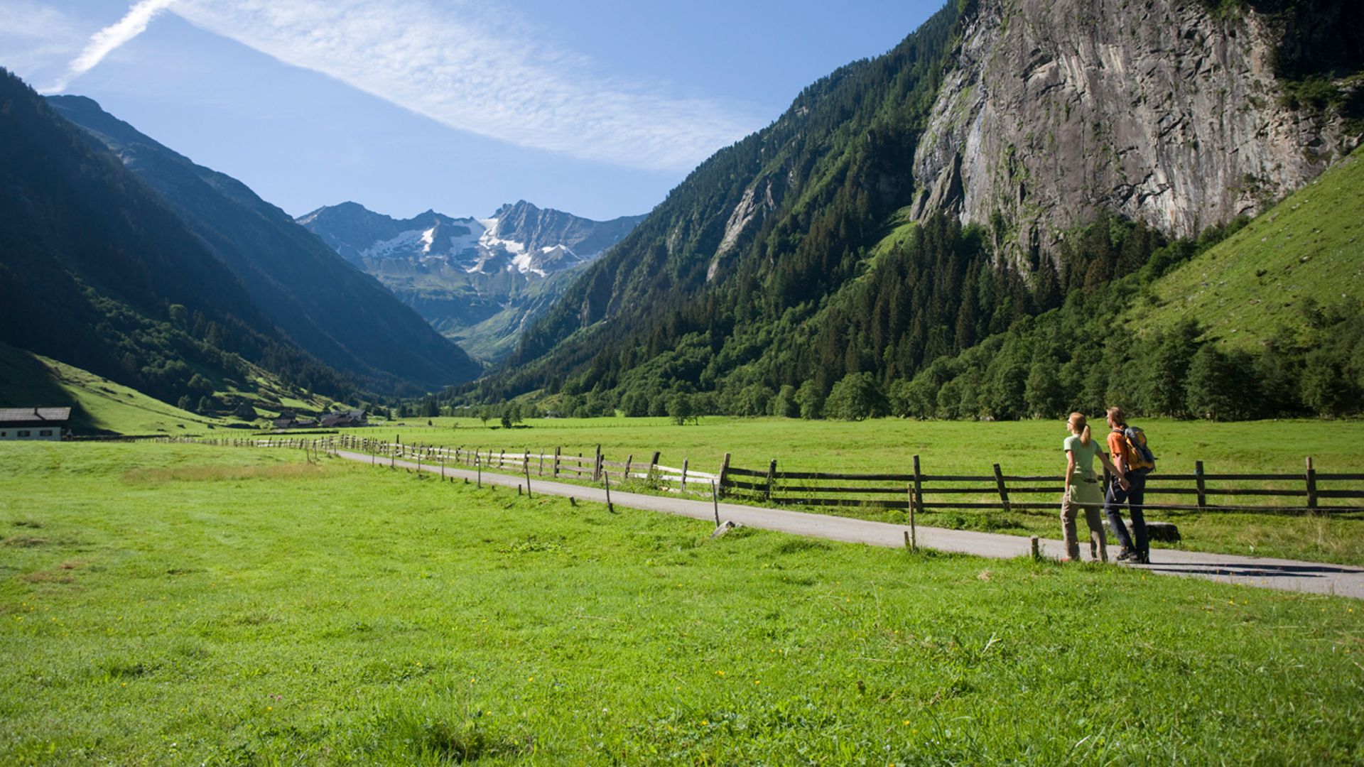 Zwei Wanderer laufen auf einem flachen Weg im Stilluptal, umgeben von weitläufigen grünen Wiesen und steilen Bergen unter einem klaren blauen Himmel.