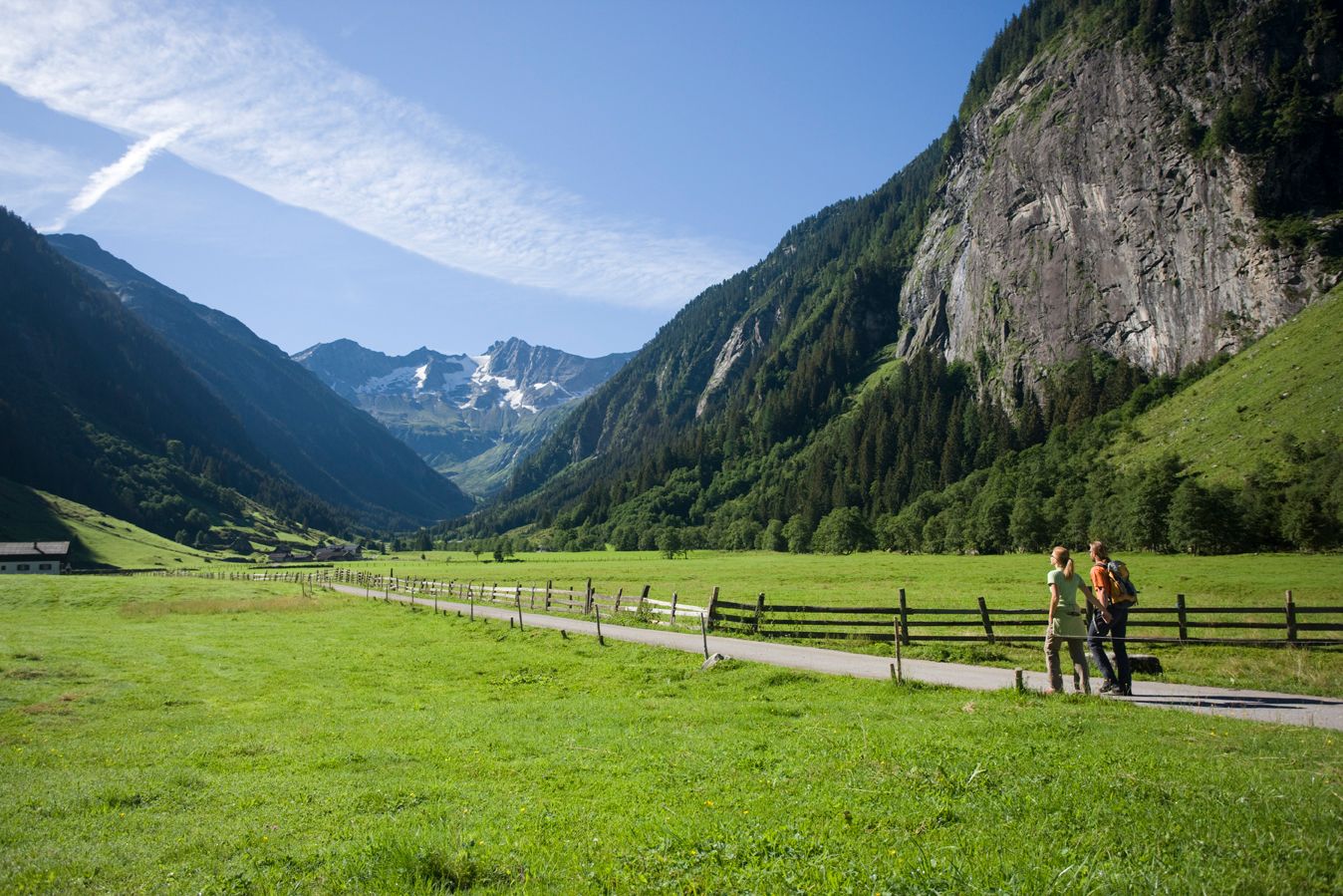 Zwei Wanderer laufen auf einem flachen Weg im Stilluptal, umgeben von weitläufigen grünen Wiesen und steilen Bergen unter einem klaren blauen Himmel.