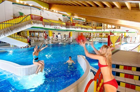 Three people play with a red ball in a colorful indoor pool. One woman stands at the edge, two others are in the water. The space features bright decorations, slides, railings, and a wooden ceiling.
