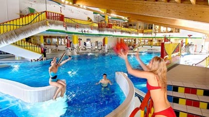 Drei Personen spielen mit einem roten Ball in einem bunten Hallenbad. Eine Frau steht am Beckenrand, zwei andere sind im Wasser. Die Umgebung ist farbenfroh gestaltet mit Rutschen, Geländern und Holzdecke.