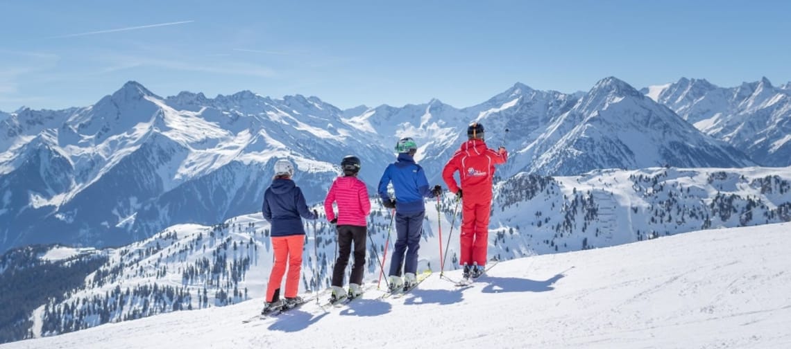 Vier Skifahrer, darunter ein Skilehrer im roten Anzug, stehen bei strahlendem Sonnenschein auf einer verschneiten Piste und blicken auf ein weites Alpen-Bergpanorama. Der Skilehrer zeigt mit dem Skistock in die Ferne.