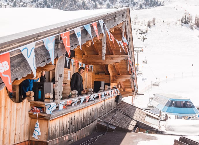 Ein DJ steht am Balkon einer Hütte am Berg, diese ist mit Fahnen dekoriert