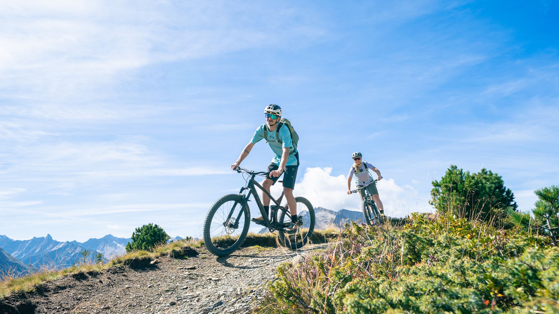 Mountain biking at Penken near Mayrhofen-Hippach: two people stand with their bikes at a viewpoint overlooking an alpine landscape with mountains under a clear blue sky.
