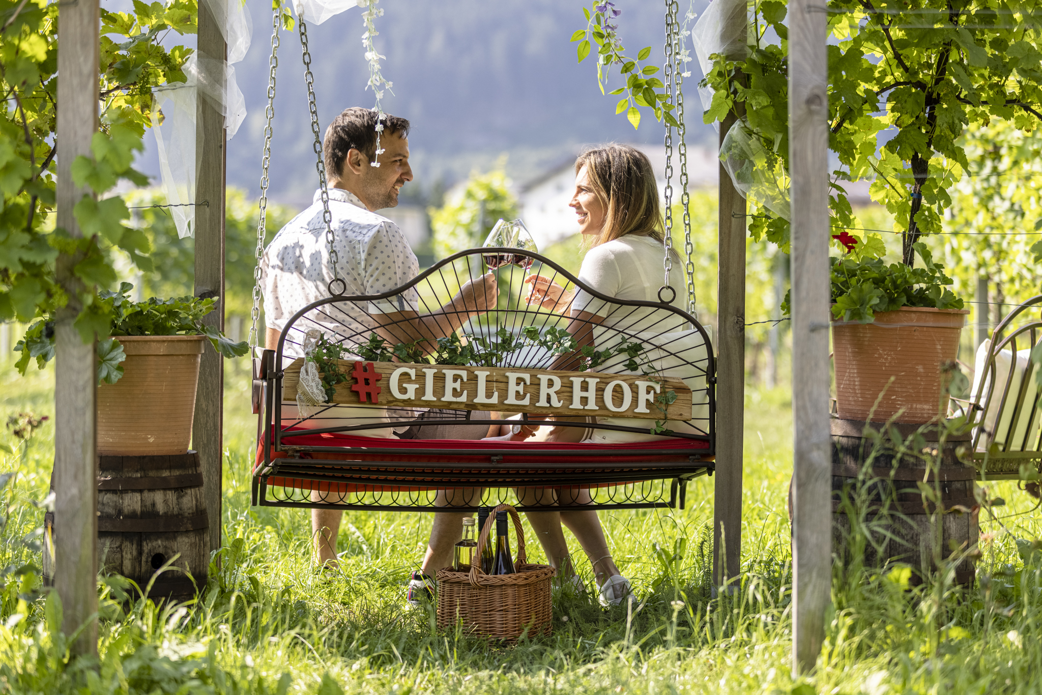 A couple sits on a swing in the garden, surrounded by plants and a wooden frame, drinking wine and smiling.