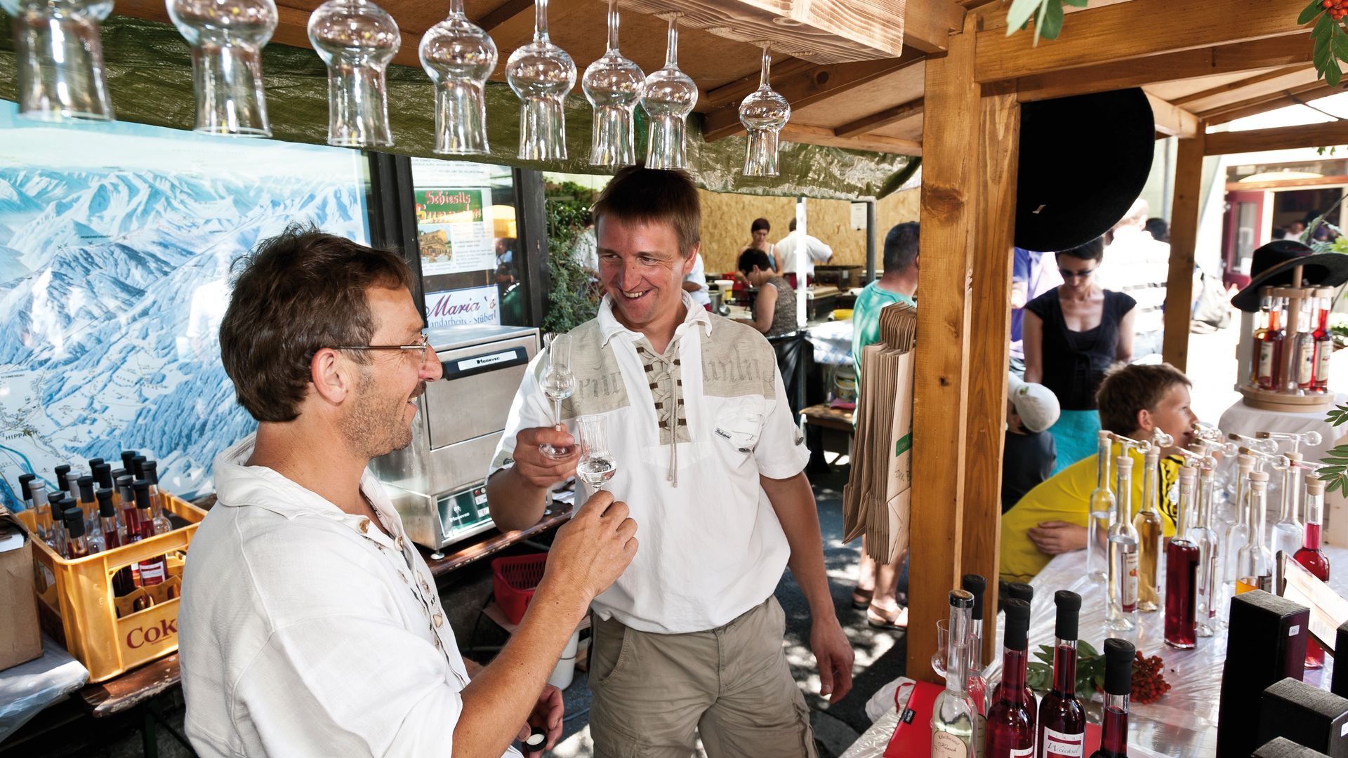 Zwei Männer stehen lachend an einer Theke und stoßen mit kleinen Gläsern an. Auf dem Tresen stehen viele Flaschen, über ihnen hängen Gläser. Holzdekor und festliche Stimmung in gemütlicher Umgebung.