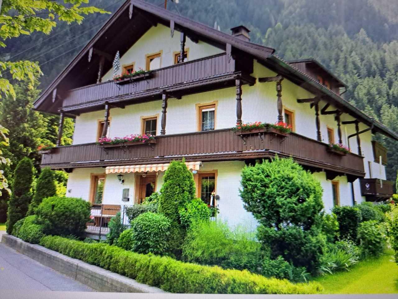 A house with wooden balconies and lush greenery, set against a forested mountain background.