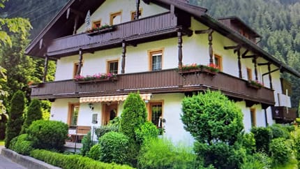 A house with wooden balconies and lush greenery, set against a forested mountain background.