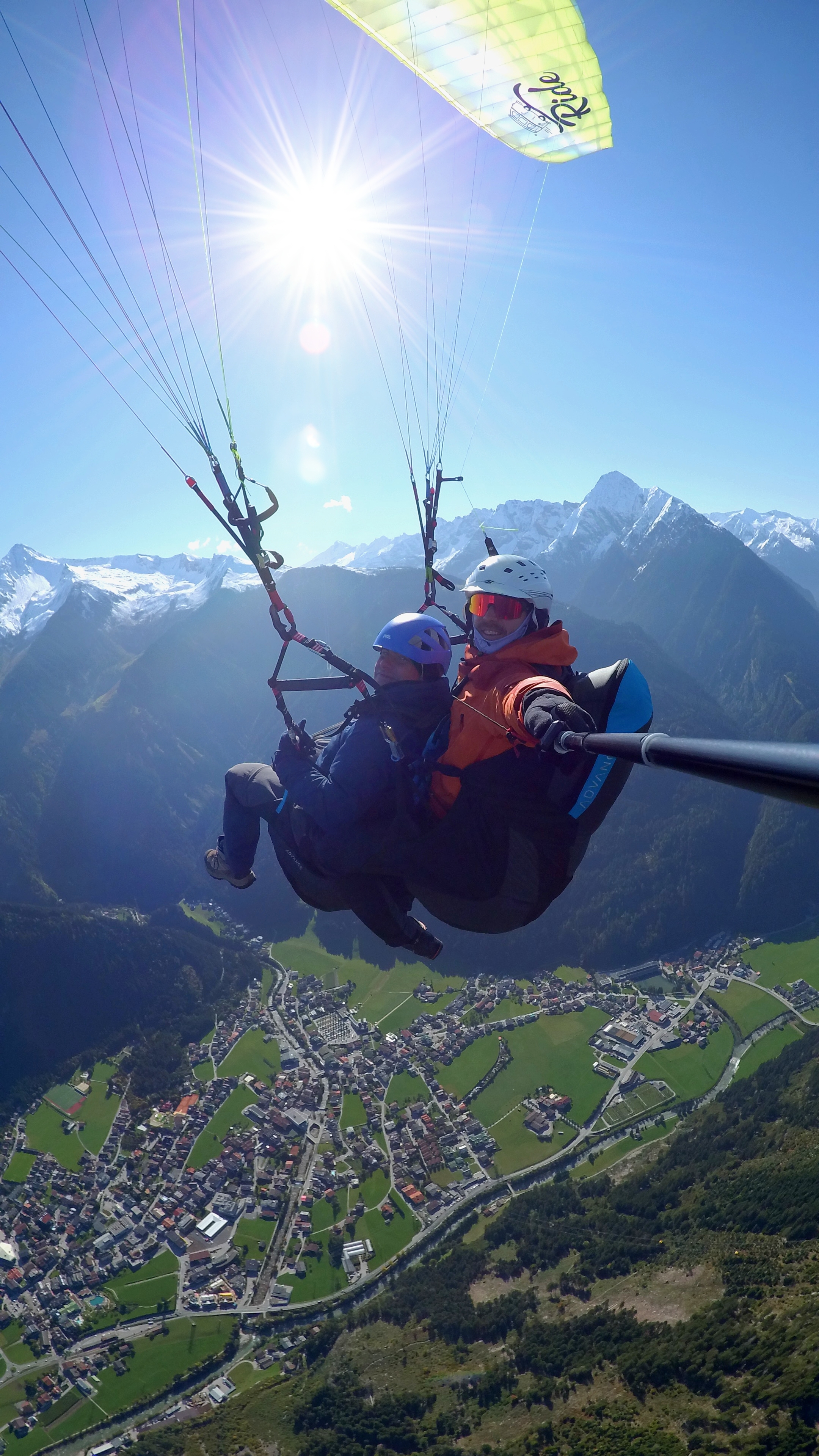Zwei Personen mit Helm auf einem Paragleitflug hoch oben am Himmel. Im Hintergrund Sonnenschein, blauer Himmel und Berge. Unter ihnen liegt das Tal mit grünen Wiesen und Häusern.