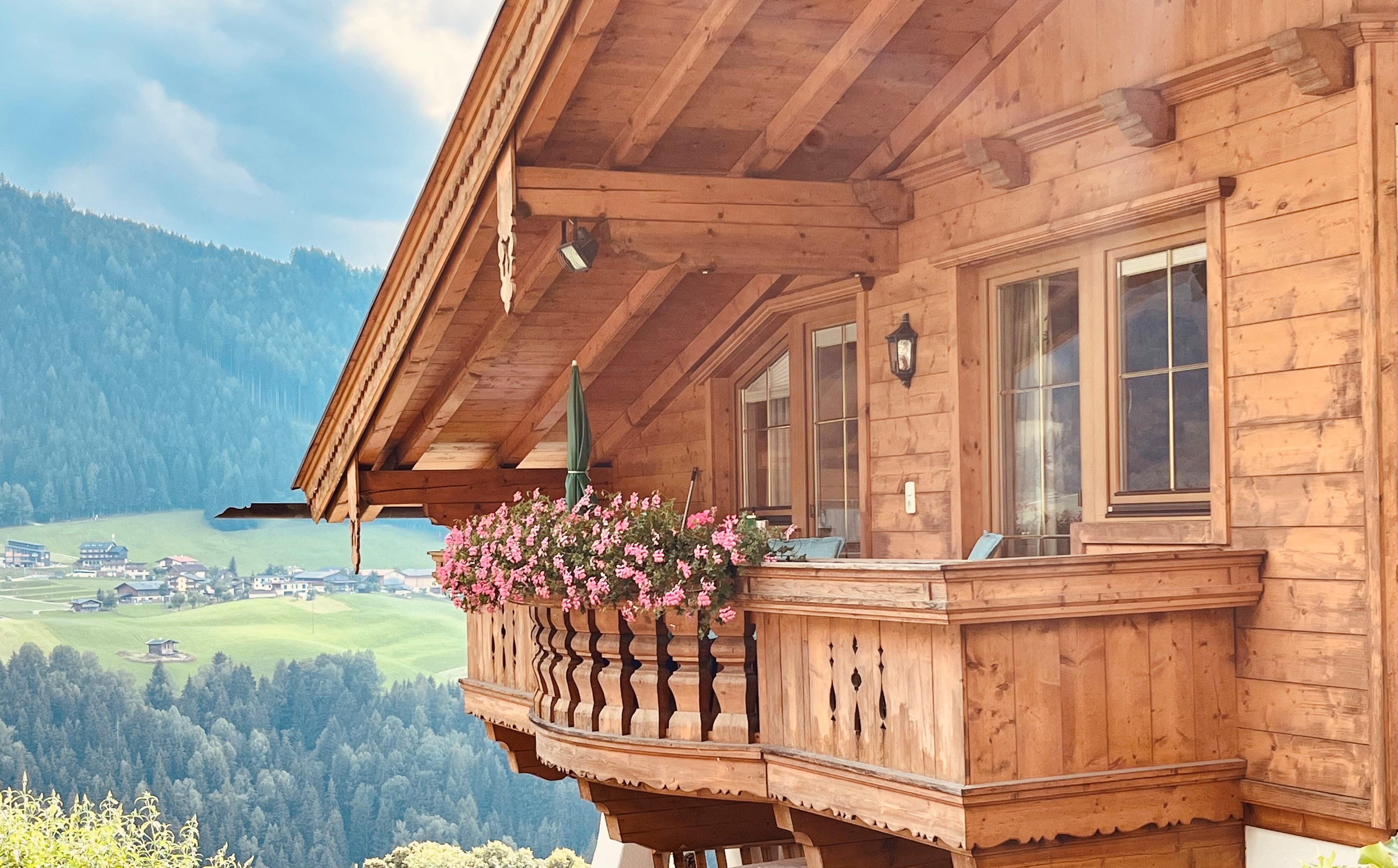 Holzhaus mit Balkon und Blumen vor Berglandschaft im Hintergrund, bei schönem Wetter.
