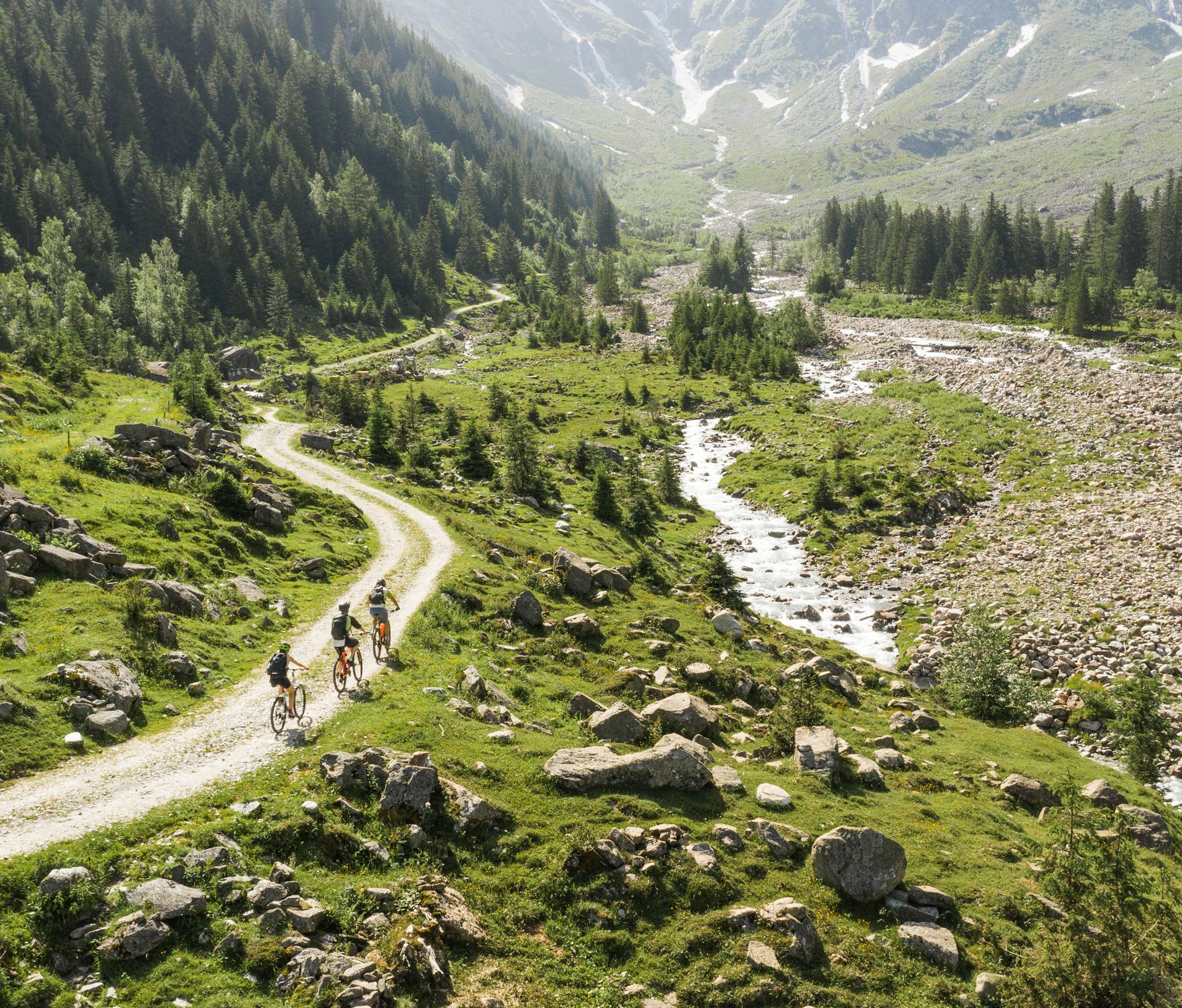 Geführte Biketour im Stilluptal: Radfahrer auf einem Weg durch grüne Berglandschaft mit Bach, Wiesen und Felsen. Sonniges Wetter in alpiner Umgebung.