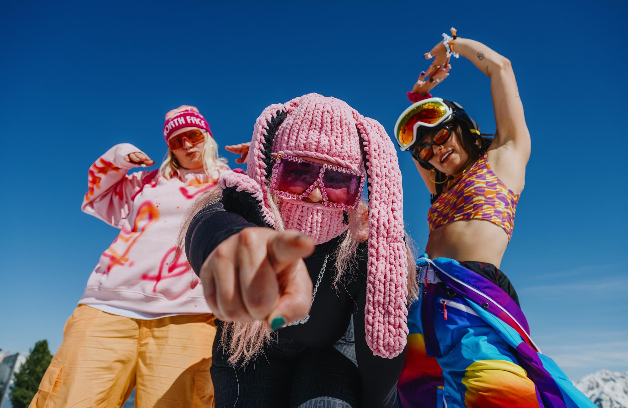 Drei junge Frauen in bunten, festivaltypischen Outfits feiern ausgelassen beim Snowbombing Festival in Mayrhofen-Hippach vor strahlend blauem Himmel.