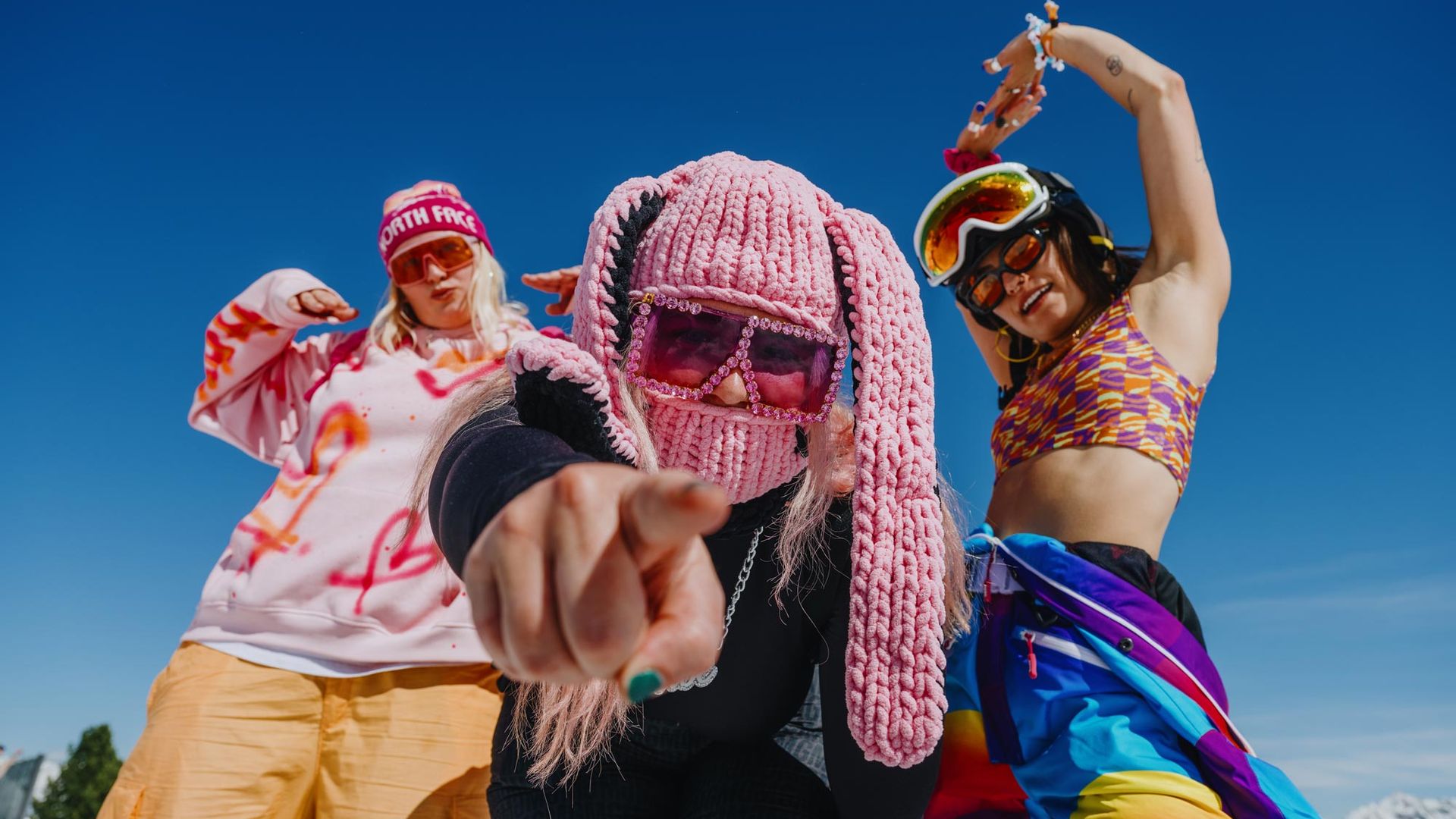 Drei junge Frauen in bunten, festivaltypischen Outfits feiern ausgelassen beim Snowbombing Festival in Mayrhofen-Hippach vor strahlend blauem Himmel.