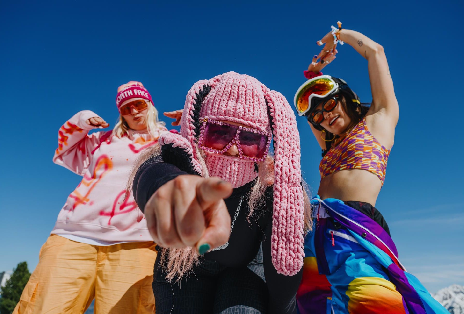 Three young women in colorful festival outfits celebrating at the Snowbombing Festival in Mayrhofen-Hippach under a bright blue sky.
