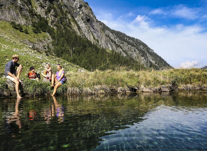 Familie sitzt am Flussufer in einer bergigen, grünen Landschaft bei schönem Himmel.