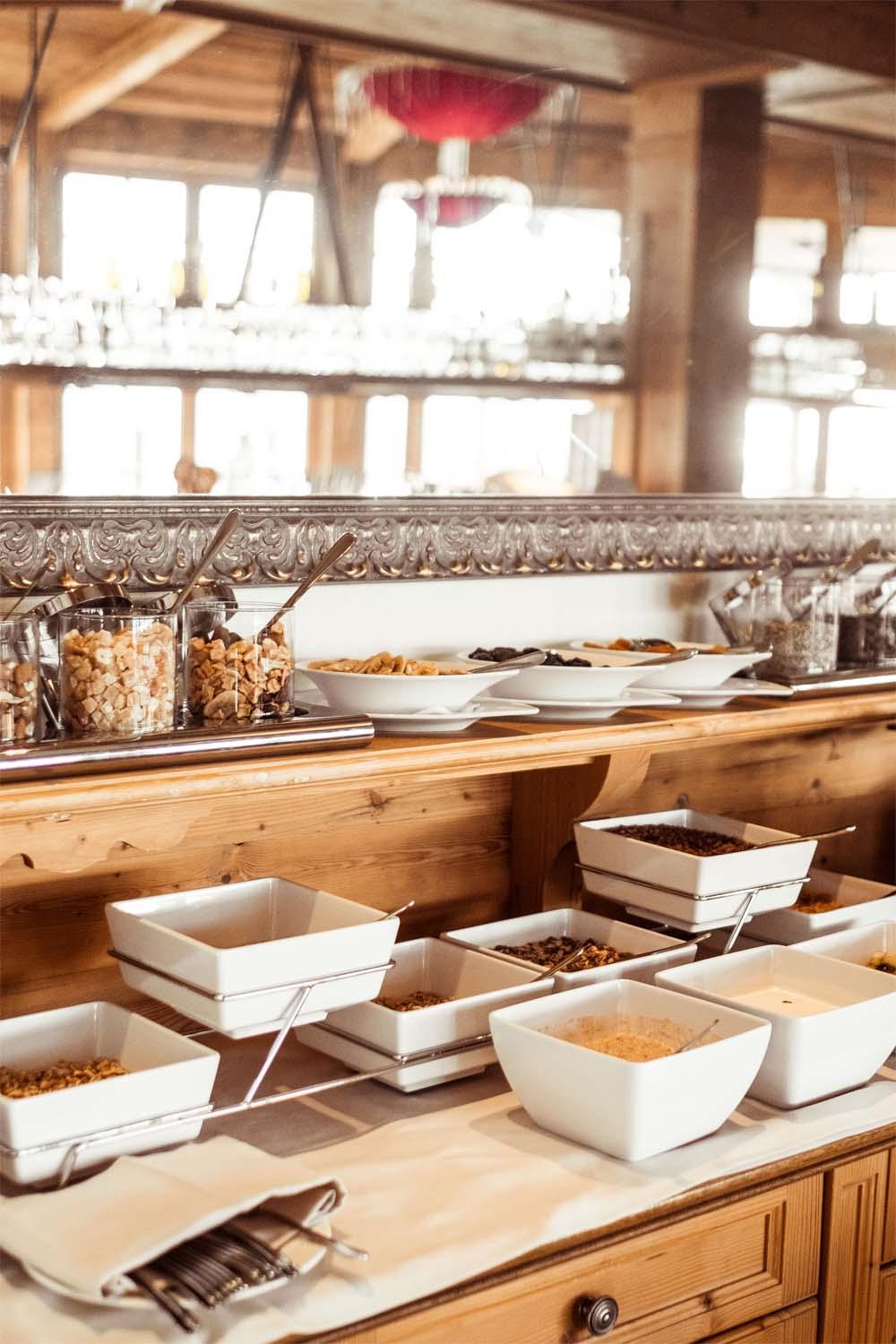 Frühstücksbuffet mit weißen Schalen voller Müsli, Nüsse und Toppings auf Holztheke, im Hintergrund Fenster und Holzdecke.