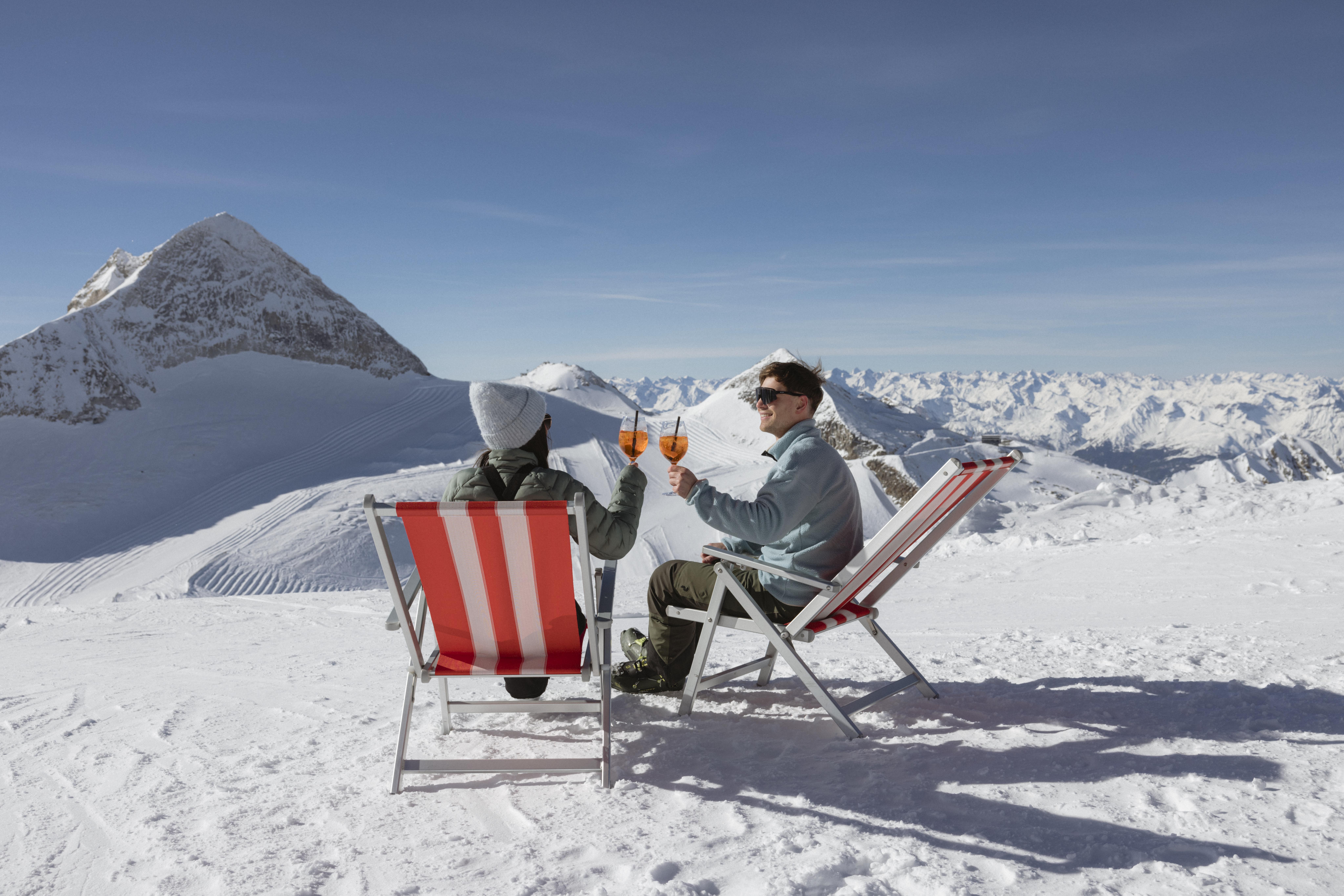 Zwei Menschen sitzen in Schneelandschaft, trinken Getränke, mit Bergen im Hintergrund, sonniges Wetter, entspannte Stimmung.