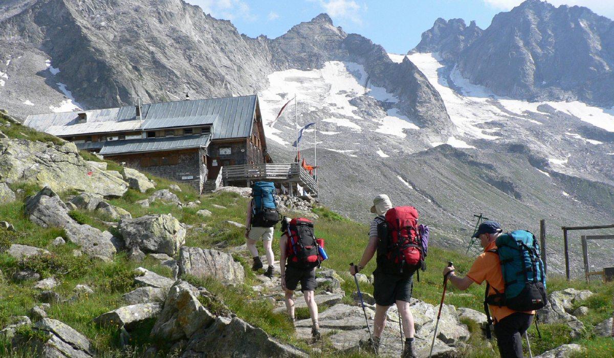 Vier Wanderer mit Rucksäcken steigen auf einem Bergpfad vor einer Berghütte, umgeben von Felsen und schneebedeckten Gipfeln.