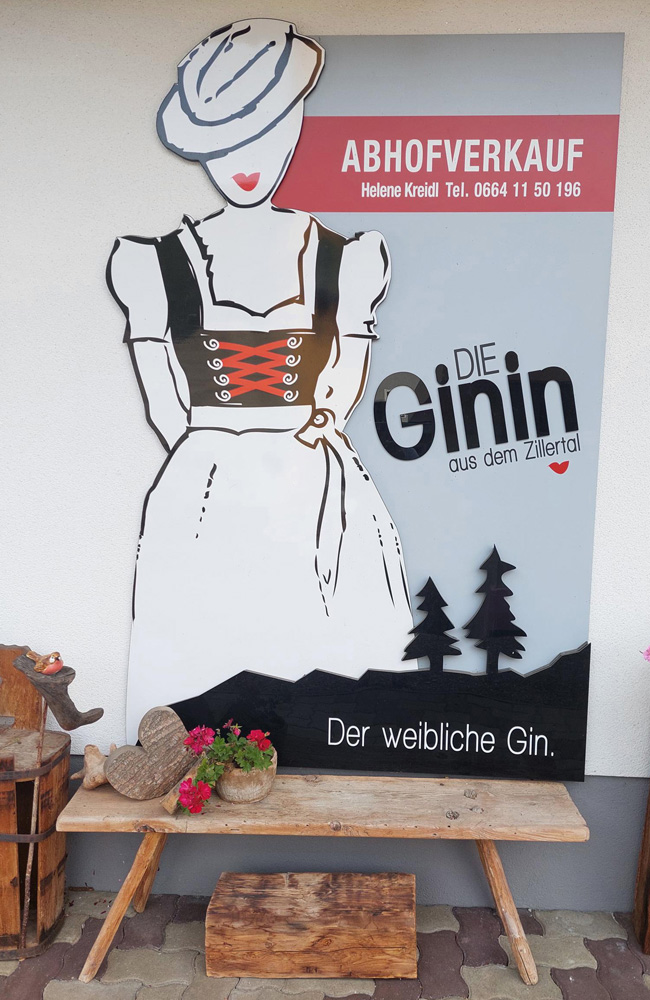 Werbetafel von „Die Ginin“ in Ramsau im Zillertal mit stilisierter Frau im Dirndl und Hinweis auf den Ab-Hof-Verkauf – Genussherbst-Partnerbetrieb der Kulinarischen Genussherbst Wochen.