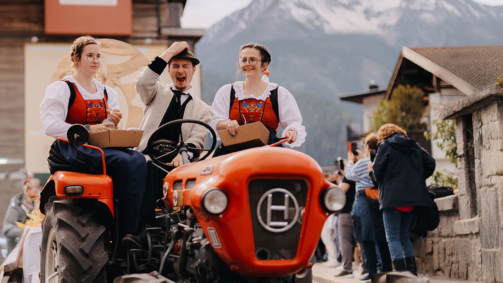 Two women in traditional dress and a man wearing a hat are riding on a red vintage tractor, laughing. People, houses, and snow-covered mountains can be seen in the background.