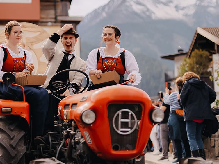 Zwei Frauen in traditioneller Tracht und ein Mann mit Hut fahren lachend auf einem roten Oldtimer-Traktor. Im Hintergrund sind Menschen, Häuser und schneebedeckte Berge zu sehen.