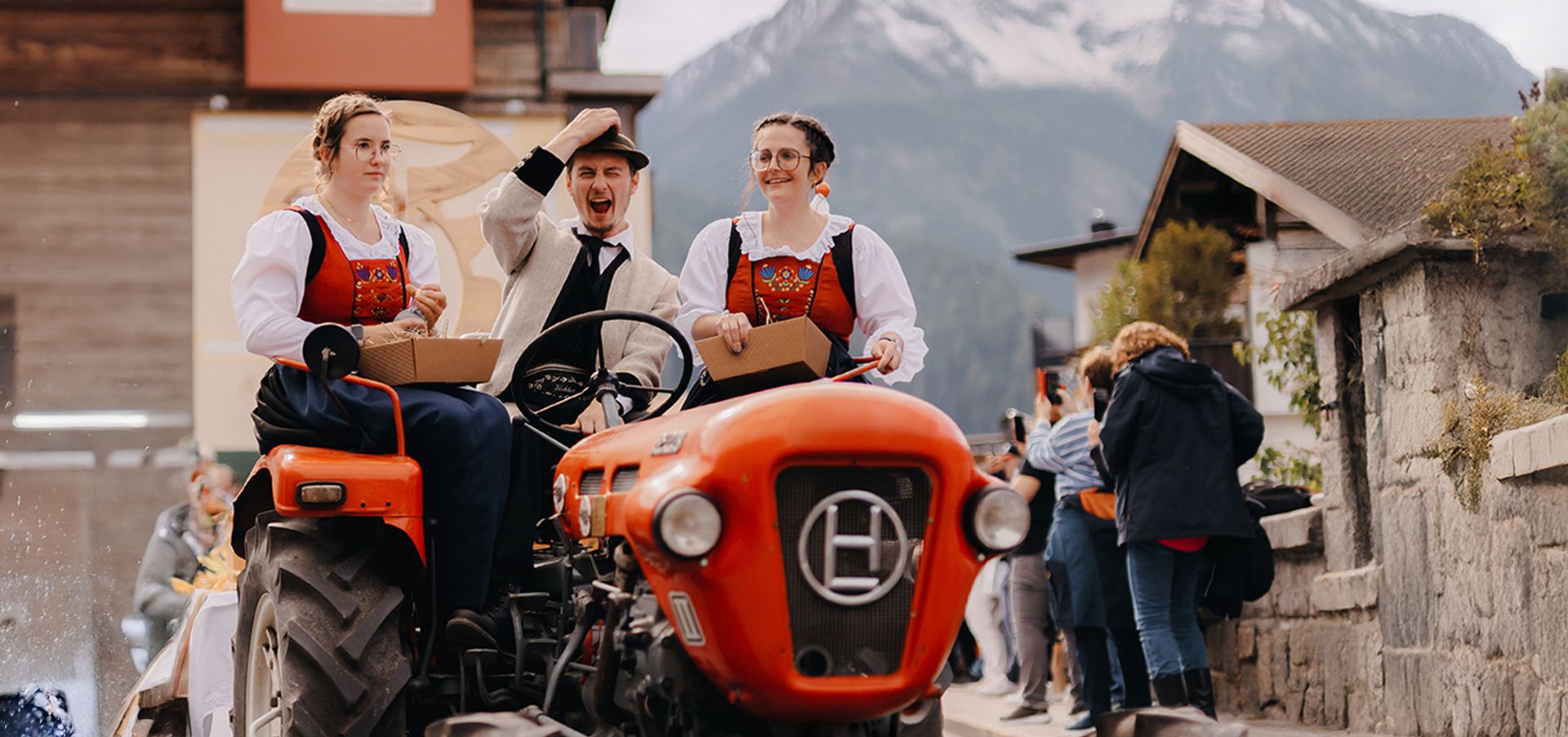 Zwei Frauen in traditioneller Tracht und ein Mann mit Hut fahren lachend auf einem roten Oldtimer-Traktor. Im Hintergrund sind Menschen, Häuser und schneebedeckte Berge zu sehen.
