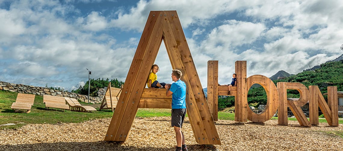 In the picture, a child is playing on a large wooden structure made up of individual letters that form the word "AHORN." A man stands nearby and watches the child. Some sun loungers can be seen in the background.