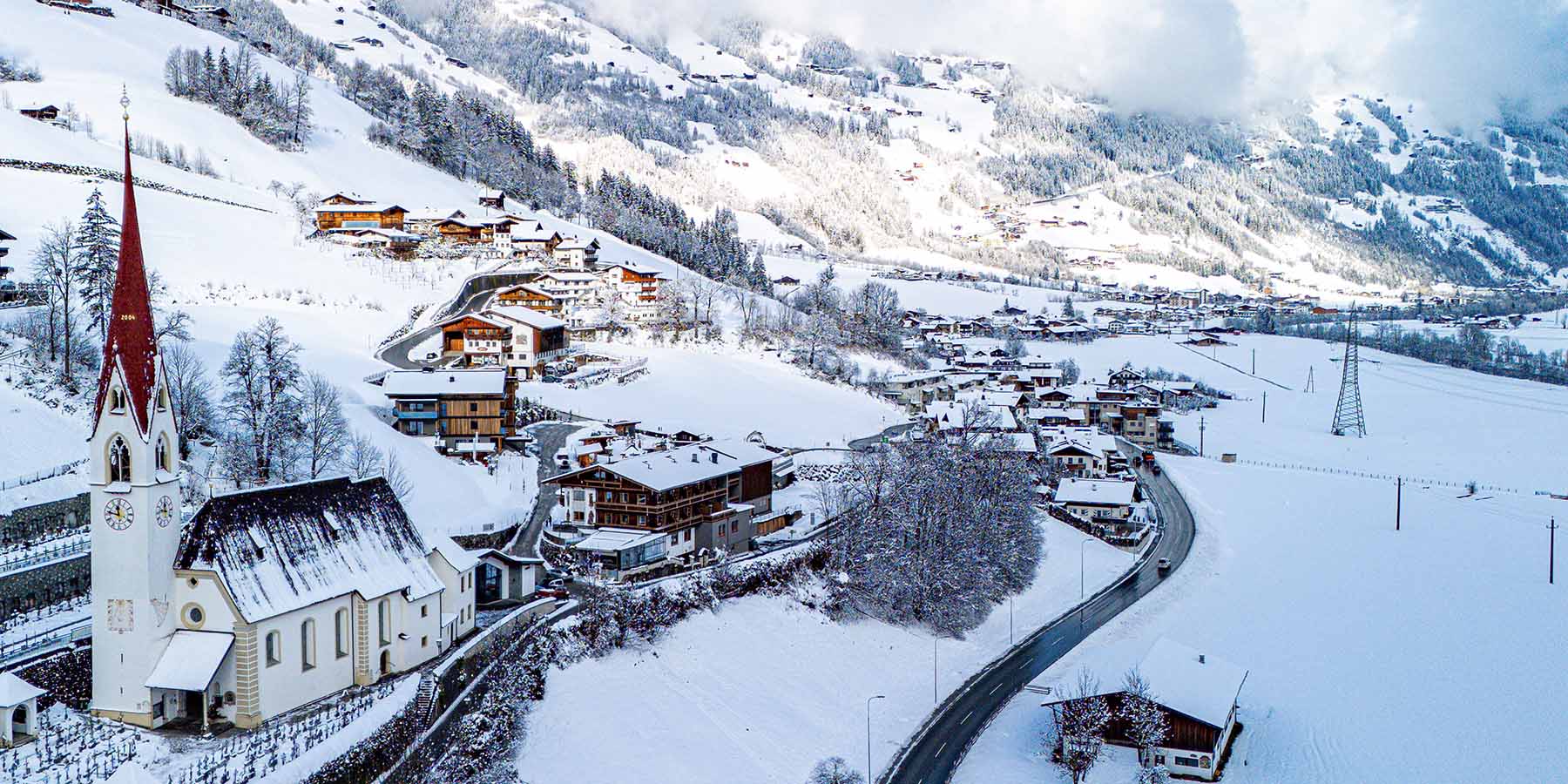 Aerial view of Hippach in the Zillertal in winter: deeply snow-covered alpine village with a distinctive red church steeple, traditional buildings with dark roofs and mighty snow-covered mountain slopes in the background. Photo: Maximilian Egger.