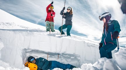 Vier Skifahrer auf Schnee, einer liegt im Schneeloch, drei stehen mit Skistöcken, alle tragen Helme und Skibrillen.