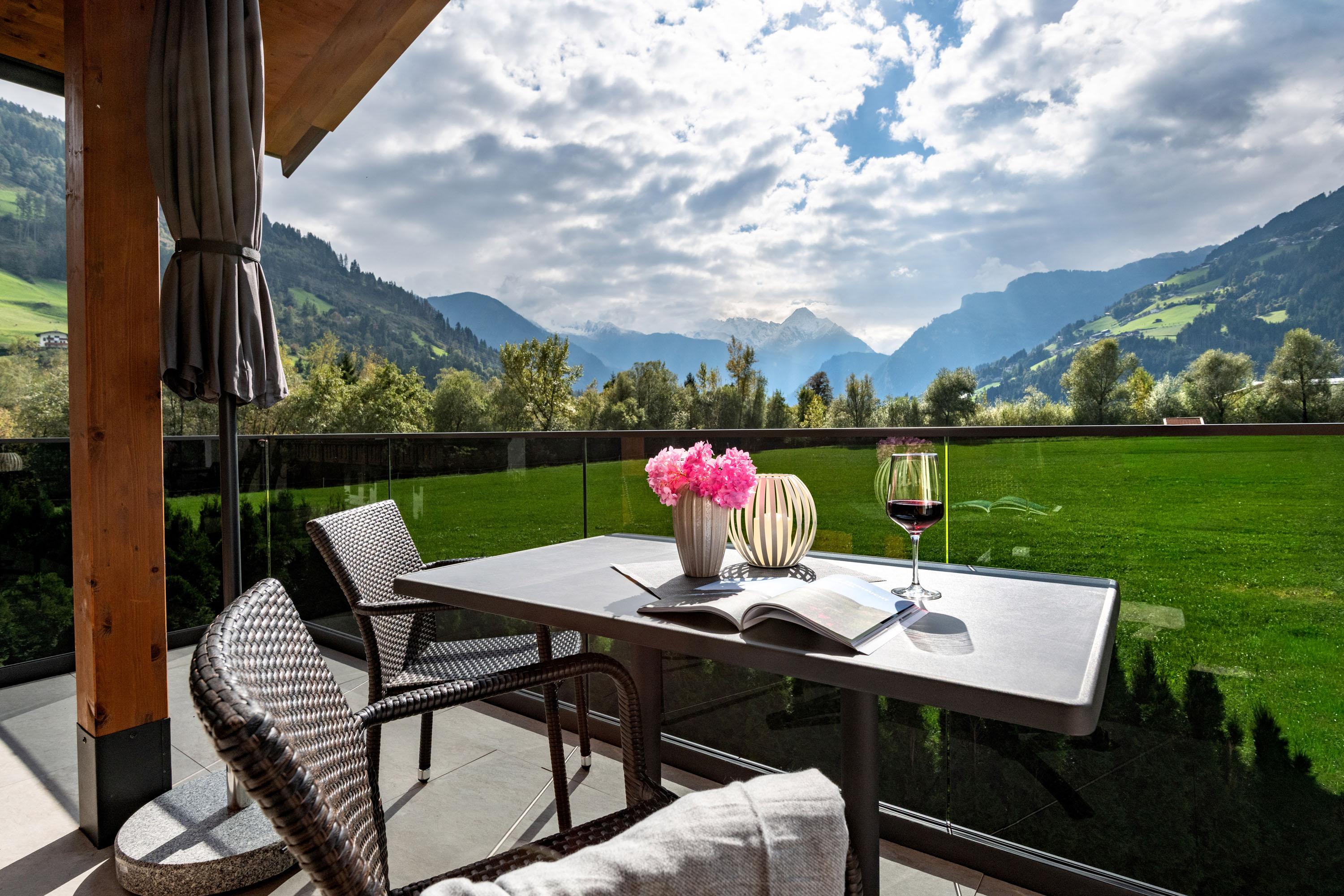 Balcony with table, flowers, wine, and mountain view under cloudy sky.