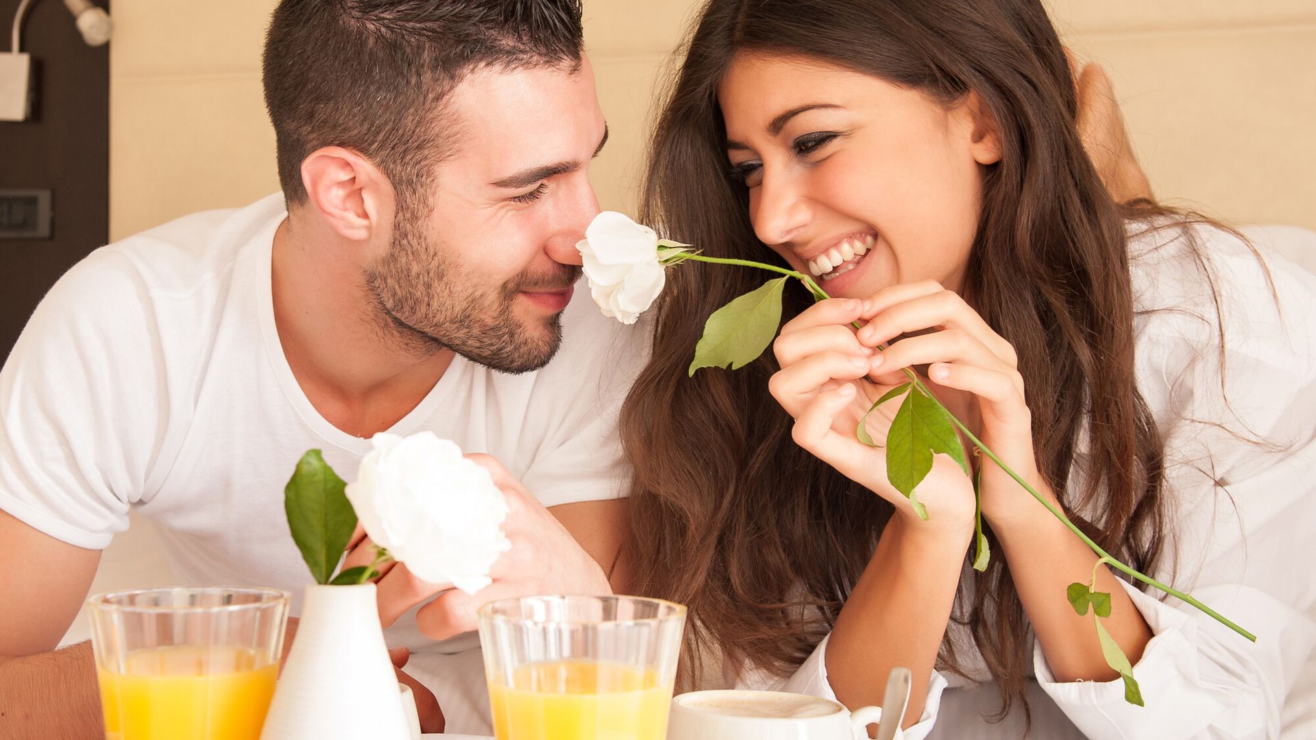 Unterkünfte in Mayrhofen-Hippach Ein junges Paar frühstückt lachend im Bett eines Hotelzimmers. Auf dem Tablett stehen Orangensaft, Kaffee und Brötchen. Die Frau hält eine weiße Rose.