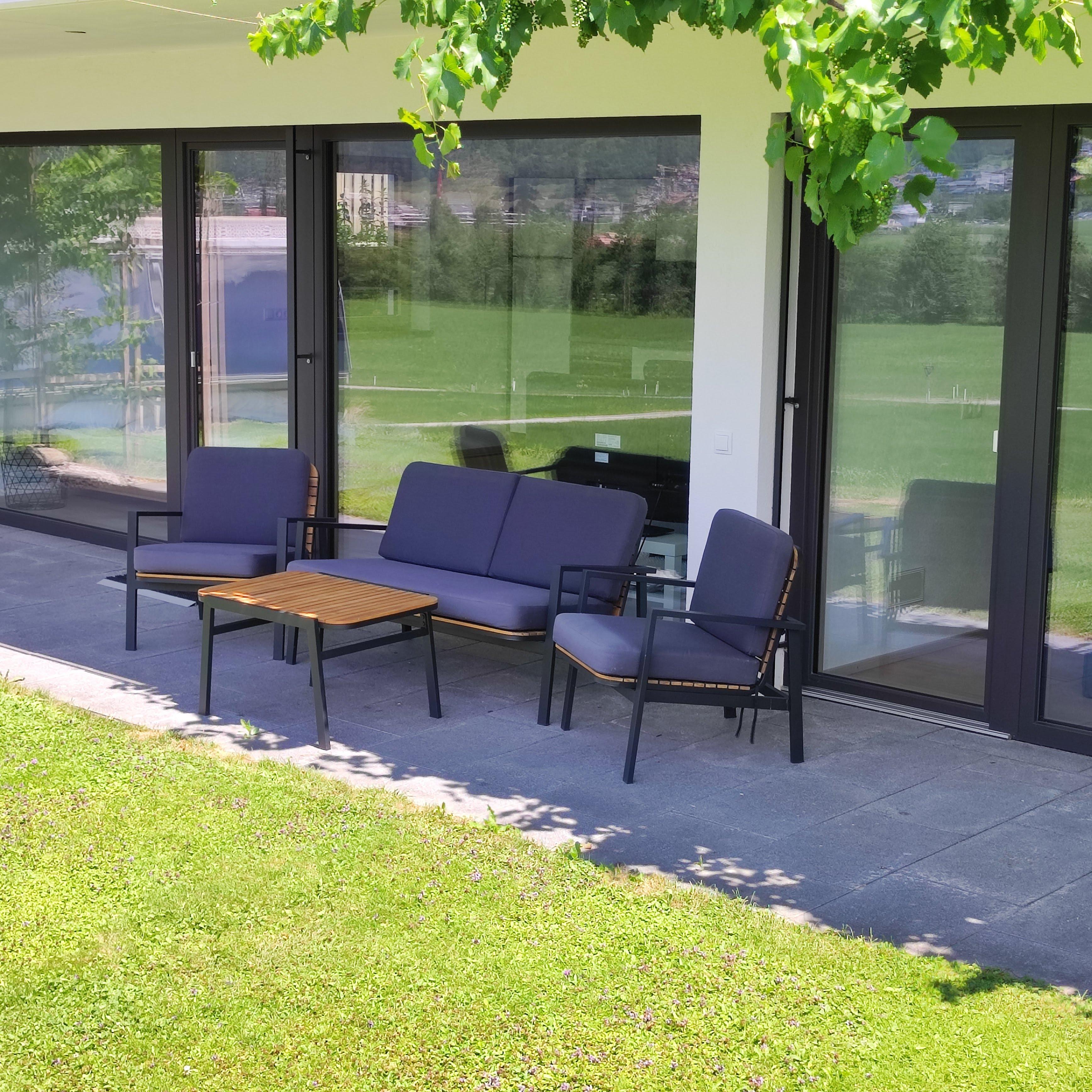 View of a modern patio with gray seating and large glass doors overlooking a green garden.