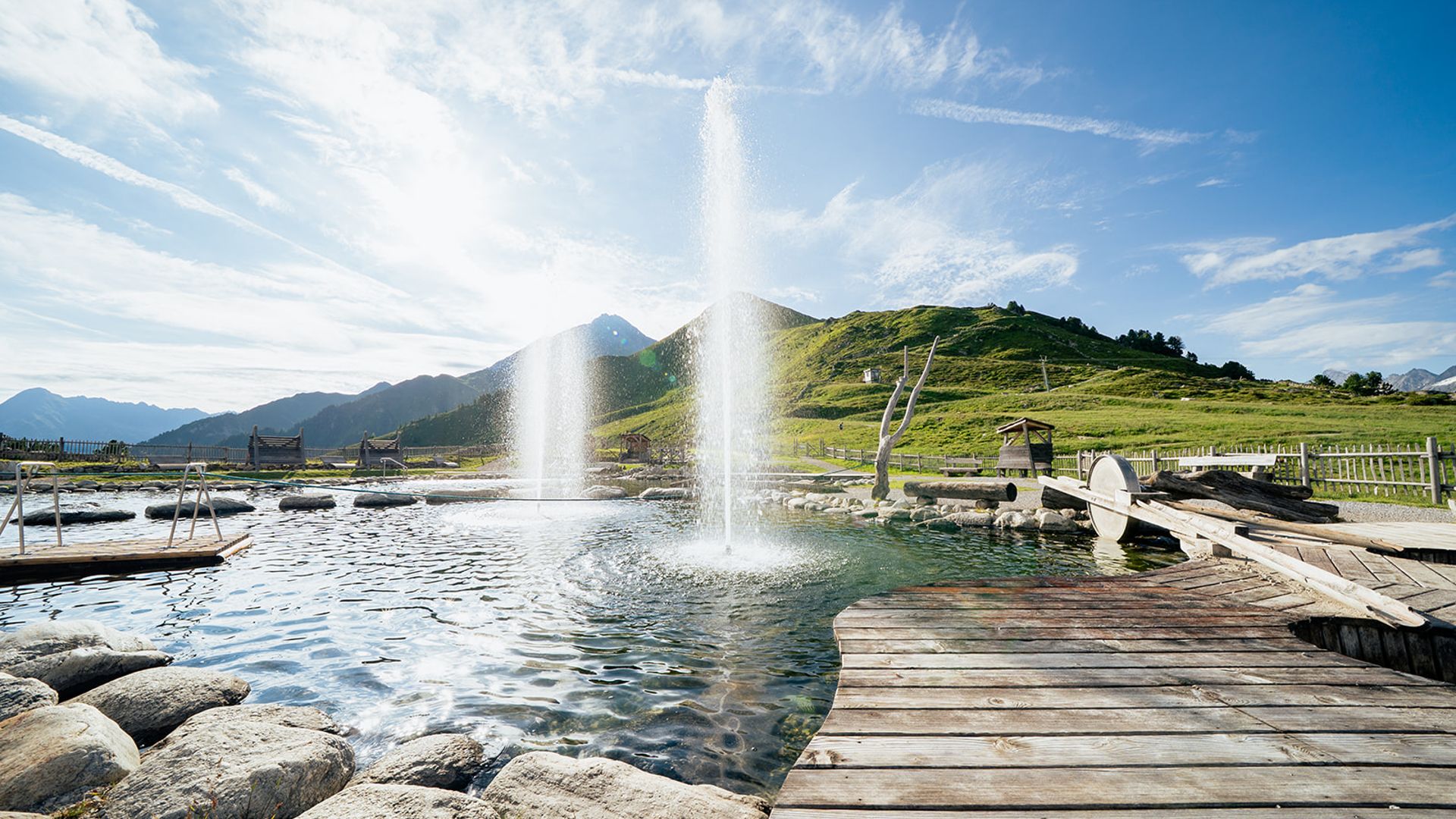 Ein idyllischer Bergsee mit klarem Wasser, umgeben von majestätischen Gipfeln. Ein geschwungener Kiesweg führt um den See. Zwei Menschen genießen die Natur – einer kniet am Ufer, eine Person in Rot sitzt entspannt.