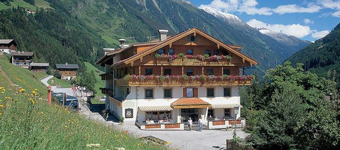 Das Bild zeigt den Gasthof Thanner von außen, mit einem Feld links daneben. Im Vordergrund stehen einige Bäume, und im Hintergrund sind Berge unter einem bewölkten Himmel zu sehen, was eine ruhige, naturnahe Atmosphäre schafft.