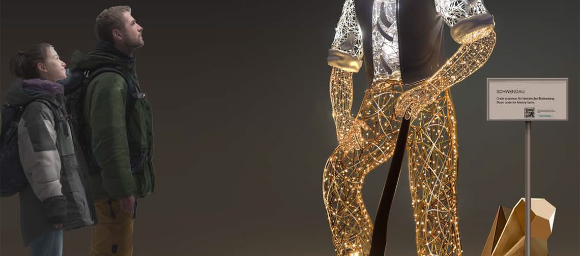 Beleuchtete Lichtskulptur in Schwendau (Tirol): stilisierte, überlebensgroße Figur mit leuchtenden Konturen und heller Kopfform steht auf abstrakter Basis; zwei Personen betrachten die Installation vor dunklem Himmel.