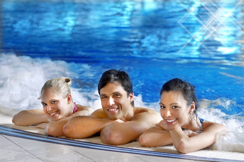 Two women and one man lie relaxed at the edge of a whirlpool. They smile at the camera. In the background, the blue pool water is bubbling gently.