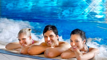 Zwei Frauen und ein Mann liegen entspannt am Beckenrand eines Whirlpools. Sie lächeln in die Kamera. Im Hintergrund ist das blaue Wasser des Pools zu sehen, es sprudelt leicht.