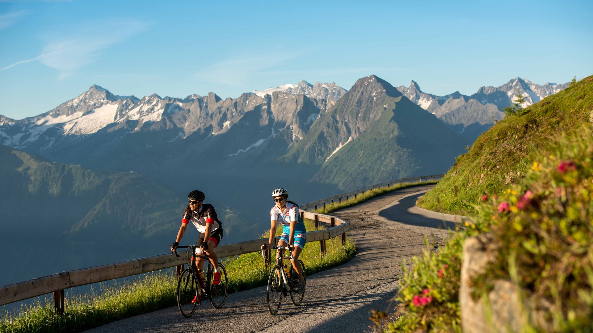 Zwei Rennradfahrer auf der Zillertaler Höhenstraße mit Blick auf die schneebedeckten Alpen. Die kurvige Panoramastraße schlängelt sich durch grüne Berglandschaft bei strahlend blauem Himmel – ideal für ambitionierte Rennradtouren.